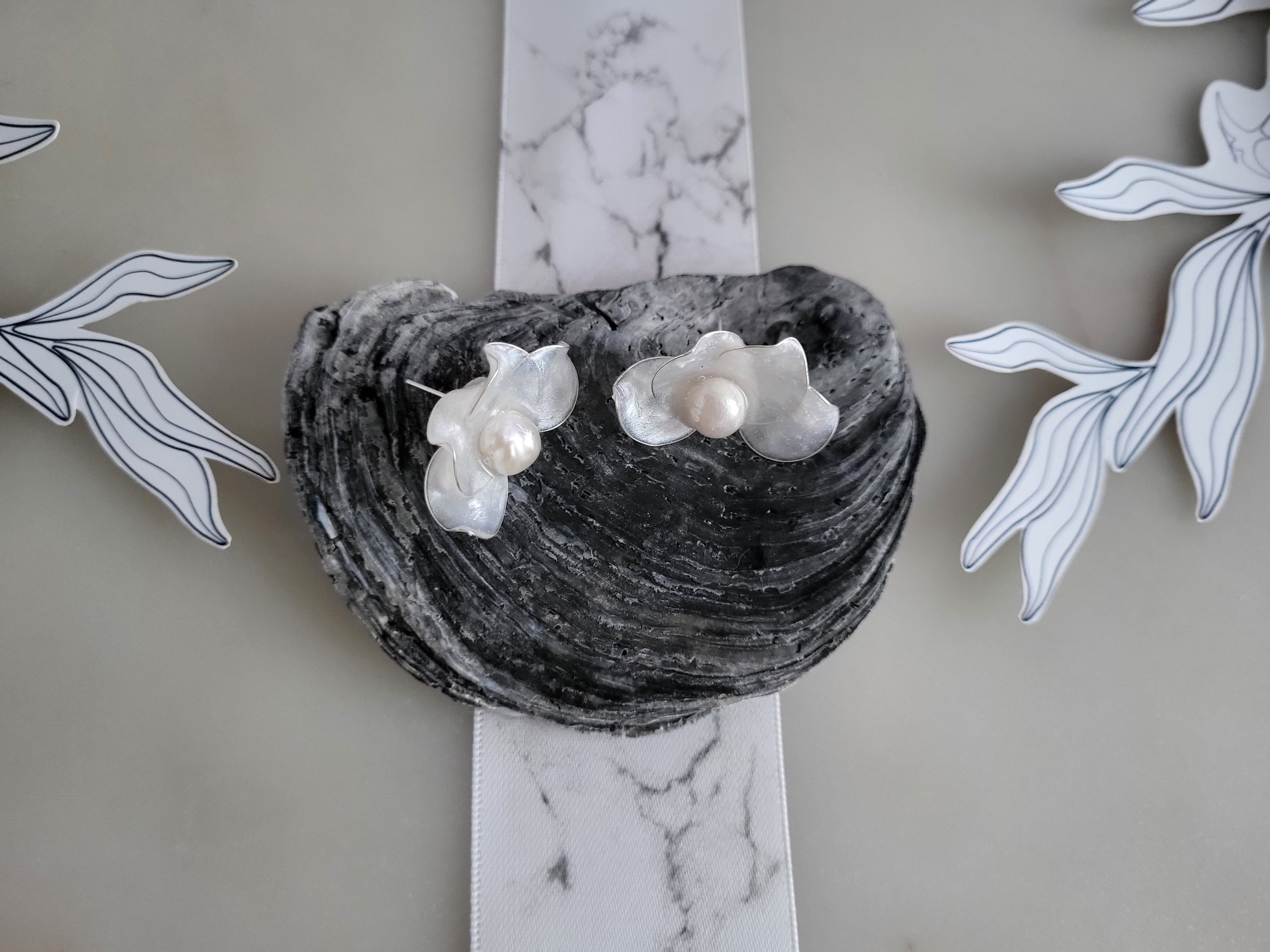 Handcrafted white water lily resin stud earrings with round natural freshwater pearls cradled in the center, displayed on a dark grey ocean shell.