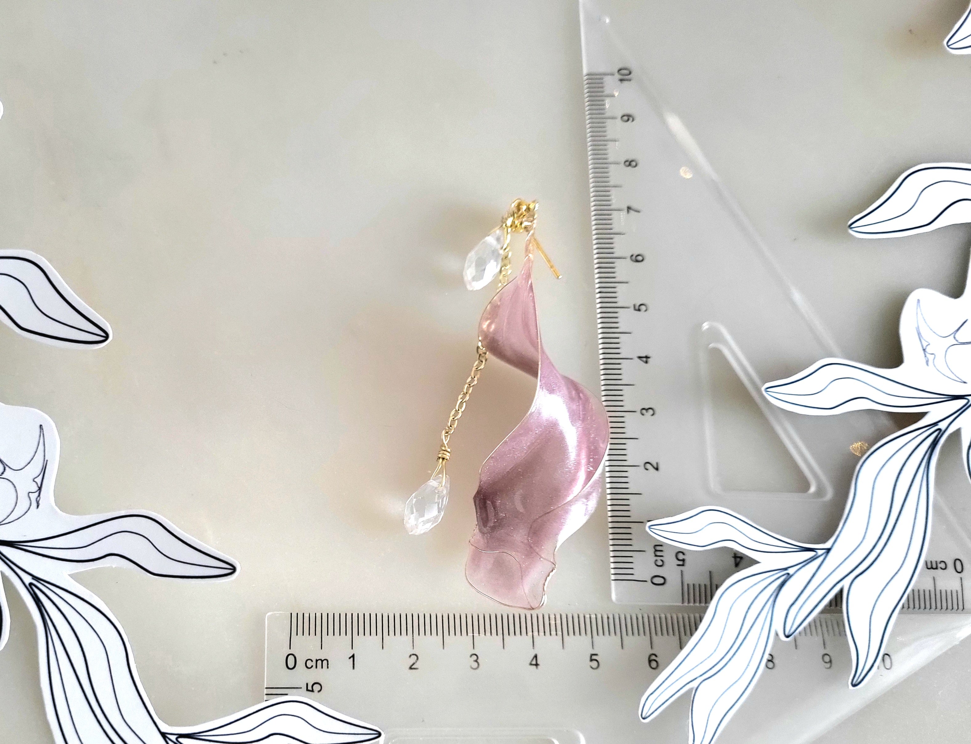 Pink leaf-shaped earring with gold accents on a white background with a ruler for scale. Handcrafted long golden wire earrings featuring pink cat eye resin flowers on a delicate chain with zirconia studs, waterdrop crystals, and raindrop dangles.